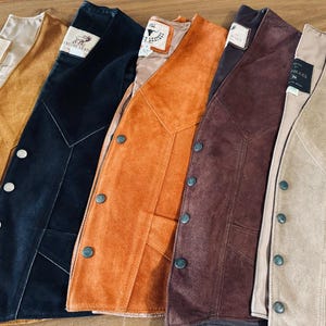 Puede incluir: Una colecci&oacute;n de chalecos de ante en varios colores, incluyendo beige, negro, naranja y marr&oacute;n. Cada chaleco tiene una parte delantera abotonada y un dise&ntilde;o cl&aacute;sico. Los chalecos est&aacute;n dispuestos uno al lado del otro, mostrando sus diferentes tonos y texturas.