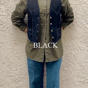 Puede incluir: Un chaleco de ante negro con botones plateados se usa sobre una camisa verde abotonada y unos vaqueros azules. La palabra "BLACK" se muestra en letras blancas en el centro de la imagen. El atuendo es informal y vintage.