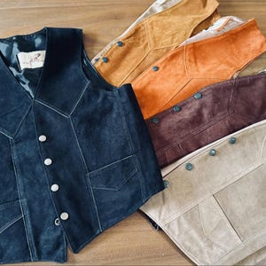Puede incluir: Una colecci&oacute;n de chalecos de ante en varios colores, incluyendo azul marino, beige, marr&oacute;n y naranja. Cada chaleco presenta una botonadura frontal, dos bolsillos delanteros y un dise&ntilde;o de cuello en V. Los chalecos se exhiben sobre una superficie de madera.