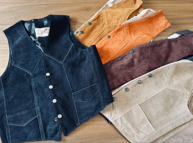 Puede incluir: Una colecci&oacute;n de chalecos de ante en varios colores, incluyendo azul marino, beige, marr&oacute;n y naranja. Cada chaleco presenta una botonadura frontal, dos bolsillos delanteros y un dise&ntilde;o de cuello en V. Los chalecos se exhiben sobre una superficie de madera.