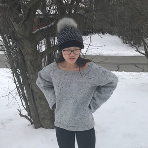 May include: A grey speckled knitted jumper with a wide neckline and long sleeves. The person is wearing a black and white patterned beanie with a grey pom-pom. The background is a snowy outdoor scene.