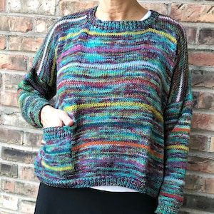 May include: A colorful, hand-knitted sweater with a round neckline and a single pocket. The sweater features horizontal stripes in various colors, including blue, green, purple, and orange. The sleeves are long, and the overall style is casual and comfortable.