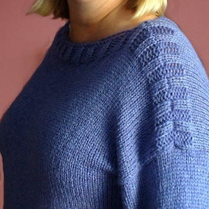 May include: A woman wearing a purple knitted jumper with a delicate lace pattern around the neckline and shoulders.