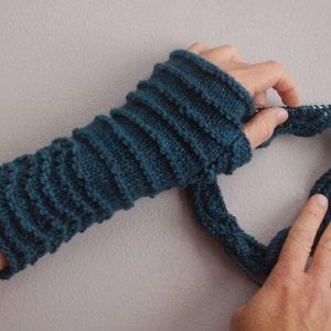 May include: A teal blue knitted fingerless glove with a ribbed pattern. The glove is being held by a person's hands, and the person is using knitting needles to create a new stitch.