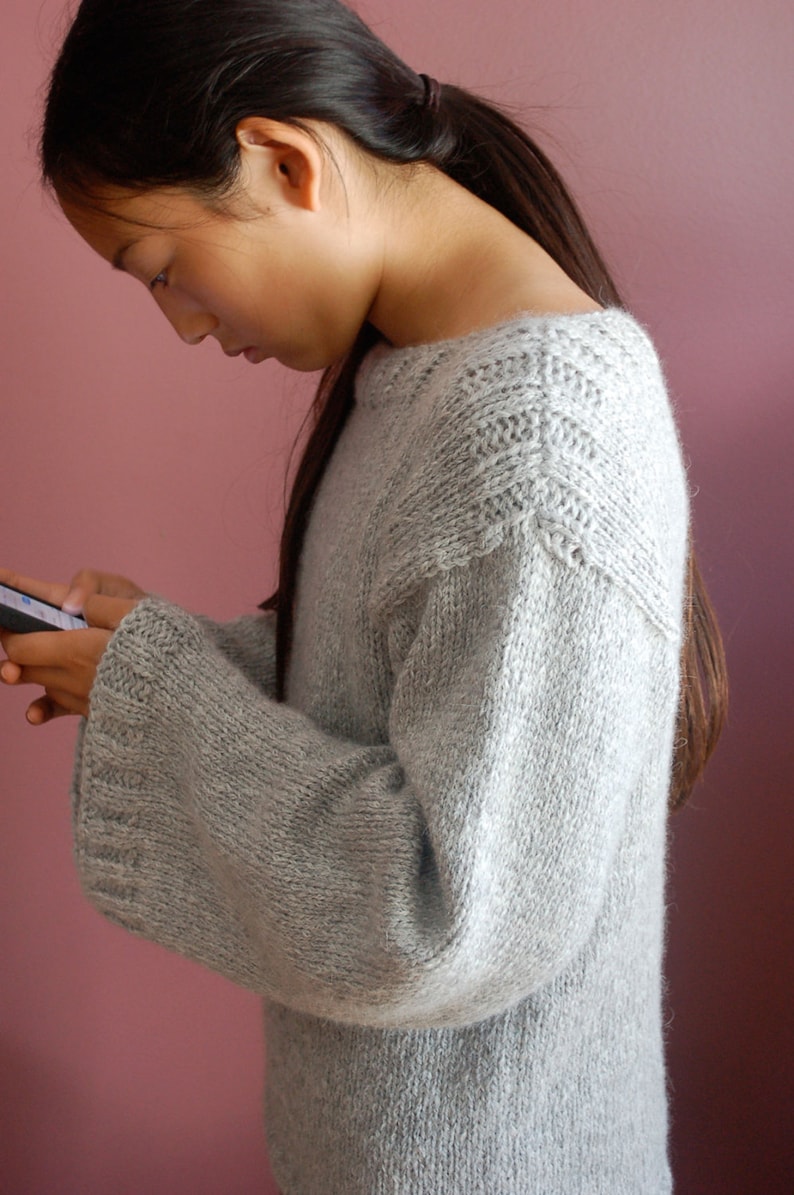 May include: A young person wearing a light gray knitted sweater with a textured pattern. The sweater has long sleeves and a round neckline.