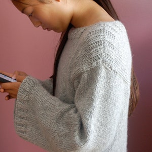 May include: A young person wearing a light gray knitted sweater with a textured pattern. The sweater has long sleeves and a round neckline.