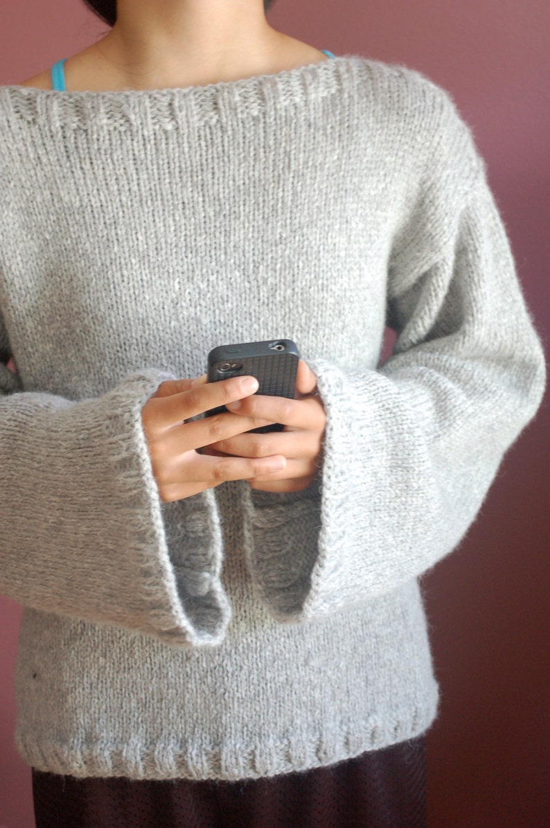 May include: A gray knitted sweater with long sleeves and a wide, boat neck. The sweater is made of a soft, fuzzy yarn and has a relaxed fit.