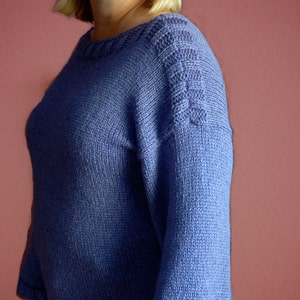 May include: A woman wearing a purple knitted sweater with a cable knit design on the shoulder.