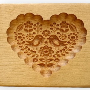 May include: A rectangular light brown wooden cookie mold featuring a deeply carved heart shape. The interior of the heart displays a detailed relief pattern of two birds facing each other, surrounded by various floral and leaf motifs.