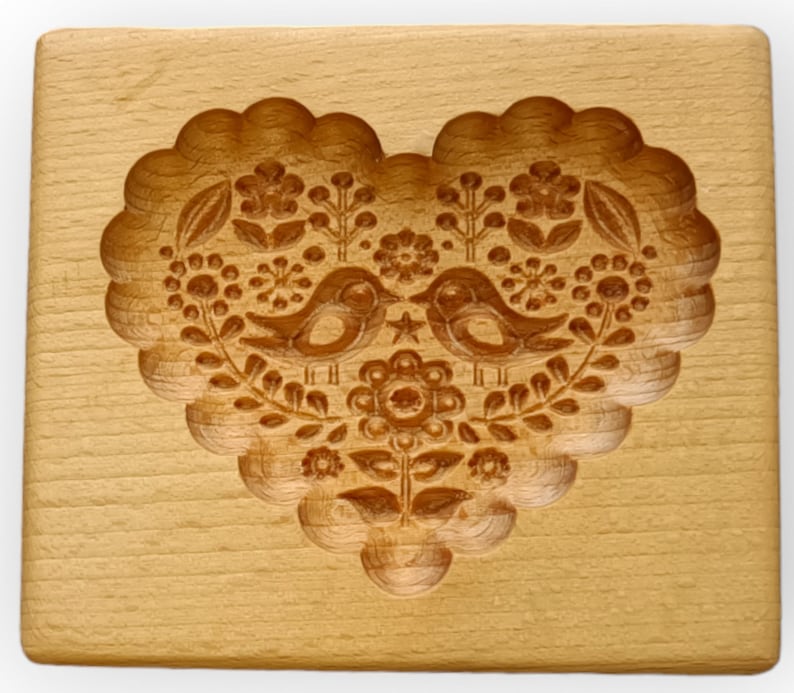 May include: A rectangular light brown wooden cookie mold featuring a deeply carved heart shape. The interior of the heart displays a detailed relief pattern of two birds facing each other, surrounded by various floral and leaf motifs.