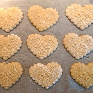 May include: Nine heart-shaped cookies arranged in a grid, each featuring an embossed design of two birds facing each other surrounded by floral patterns. The cookies have a scalloped edge and a light beige color, showing intricate textures.