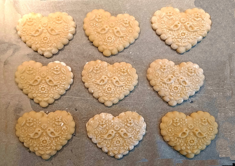 May include: Nine heart-shaped cookies arranged in a grid, each featuring an embossed design of two birds facing each other surrounded by floral patterns. The cookies have a scalloped edge and a light beige color, showing intricate textures.