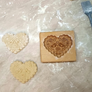 May include: A rectangular wooden cookie mold with a heart-shaped cavity featuring an embossed design of two birds surrounded by flowers, accompanied by two heart-shaped cookies with the same pattern and a grey silicone pastry brush.
