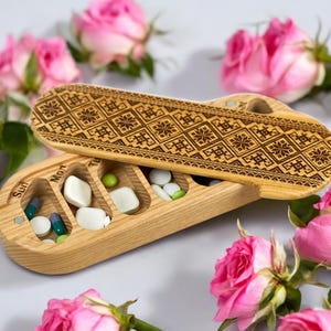 Puede incluir: Una caja de pastillas de madera con una tapa que presenta un patrón geométrico. La caja está dividida en siete compartimentos etiquetados con los días de la semana: Dom, Lun, Mar, Mié, Jue, Vie, Sáb. La caja está abierta y contiene varias pastillas.