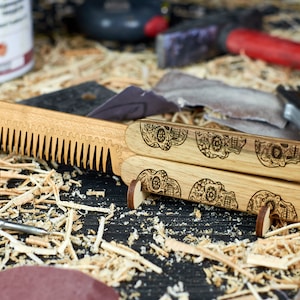 May include: A wooden comb with a skull design on the handle. The comb is folded in half and is sitting on a wooden surface with wood shavings.