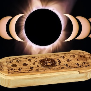 Peut inclure: Une boîte en bois avec un motif de phases lunaires gravé sur le dessus. La boîte est fermée et a une finition en bois naturel. L'arrière-plan est une image floue d'une éclipse solaire.