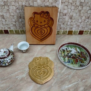Puede incluir: Un cortador de galletas de madera con forma de Hello Kitty sosteniendo un coraz&oacute;n. El cortador est&aacute; sobre una superficie de madera. Hay una peque&ntilde;a tetera blanca, una peque&ntilde;a taza de t&eacute; blanca y un peque&ntilde;o plato blanco con un dise&ntilde;o colorido.