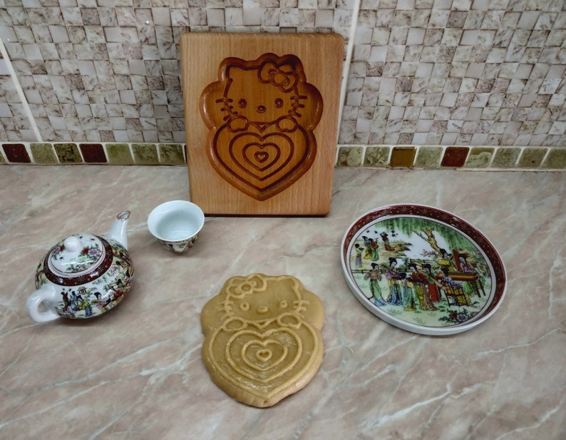 Puede incluir: Un cortador de galletas de madera con forma de Hello Kitty sosteniendo un coraz&oacute;n. El cortador est&aacute; sobre una superficie de madera. Hay una peque&ntilde;a tetera blanca, una peque&ntilde;a taza de t&eacute; blanca y un peque&ntilde;o plato blanco con un dise&ntilde;o colorido.