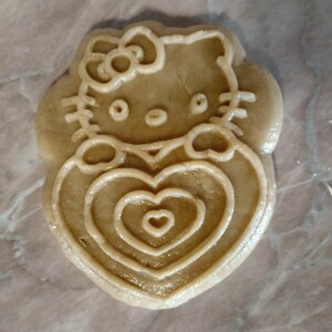 Puede incluir: Una galleta marr&oacute;n con un dise&ntilde;o de Hello Kitty. La galleta tiene forma de coraz&oacute;n con tres c&iacute;rculos conc&eacute;ntricos en su interior.
