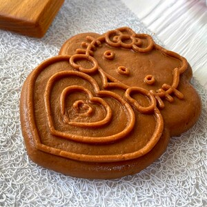 Puede incluir: Una galleta de coraz&oacute;n marr&oacute;n con un dise&ntilde;o de Hello Kitty en relieve. La galleta est&aacute; decorada con un lazo y un coraz&oacute;n.