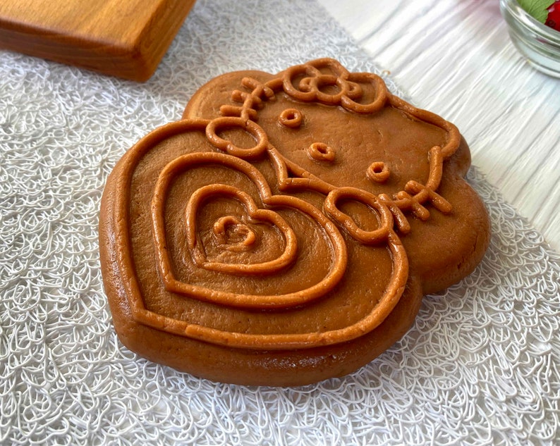 Puede incluir: Una galleta de coraz&oacute;n marr&oacute;n con un dise&ntilde;o de Hello Kitty en relieve. La galleta est&aacute; decorada con un lazo y un coraz&oacute;n.
