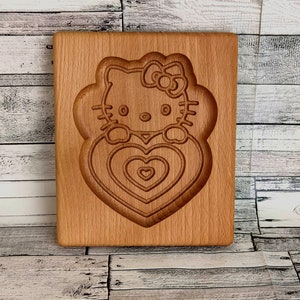 Puede incluir: Un cortador de galletas de madera con forma de Hello Kitty sosteniendo un coraz&oacute;n. El cortador tiene un dise&ntilde;o detallado con m&uacute;ltiples formas de coraz&oacute;n.