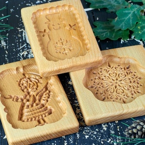 May include: Three wooden cookie cutters, one shaped like a snowman, one shaped like an angel, and one shaped like a snowflake. The cutters are on a dark wood surface.