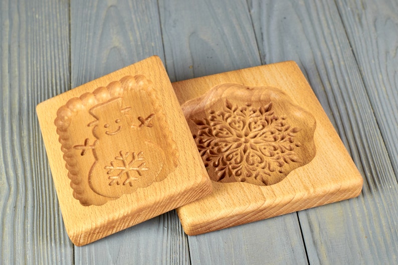 May include: Two wooden cookie cutters, one with a snowflake design and the other with a snowman design. The cutters are made of light brown wood and have a smooth finish.