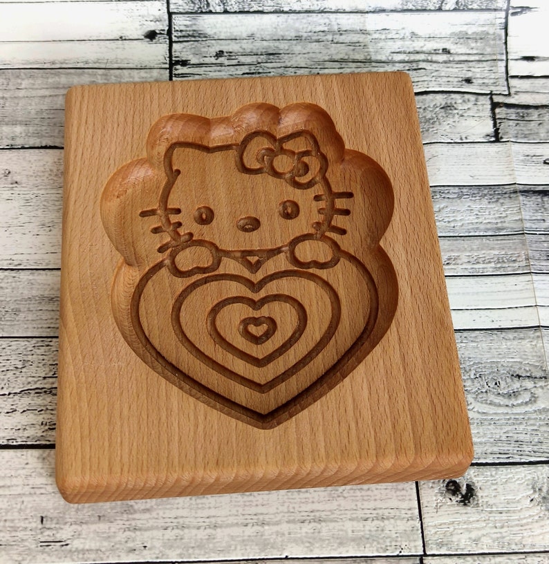 Puede incluir: Cortador de galletas de madera con un dise&ntilde;o de Hello Kitty sosteniendo un coraz&oacute;n. El cortador est&aacute; hecho de madera marr&oacute;n claro y tiene un acabado liso.