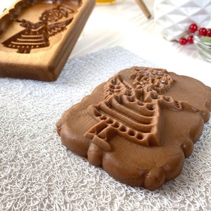 May include: A brown gingerbread cookie with an embossed design of an angel. The cookie is sitting on a white lace doily.