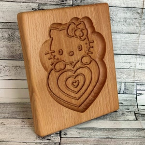 Puede incluir: Cortador de galletas de madera con un dise&ntilde;o tallado de Hello Kitty sosteniendo un coraz&oacute;n. El coraz&oacute;n tiene c&iacute;rculos conc&eacute;ntricos de corazones m&aacute;s peque&ntilde;os.