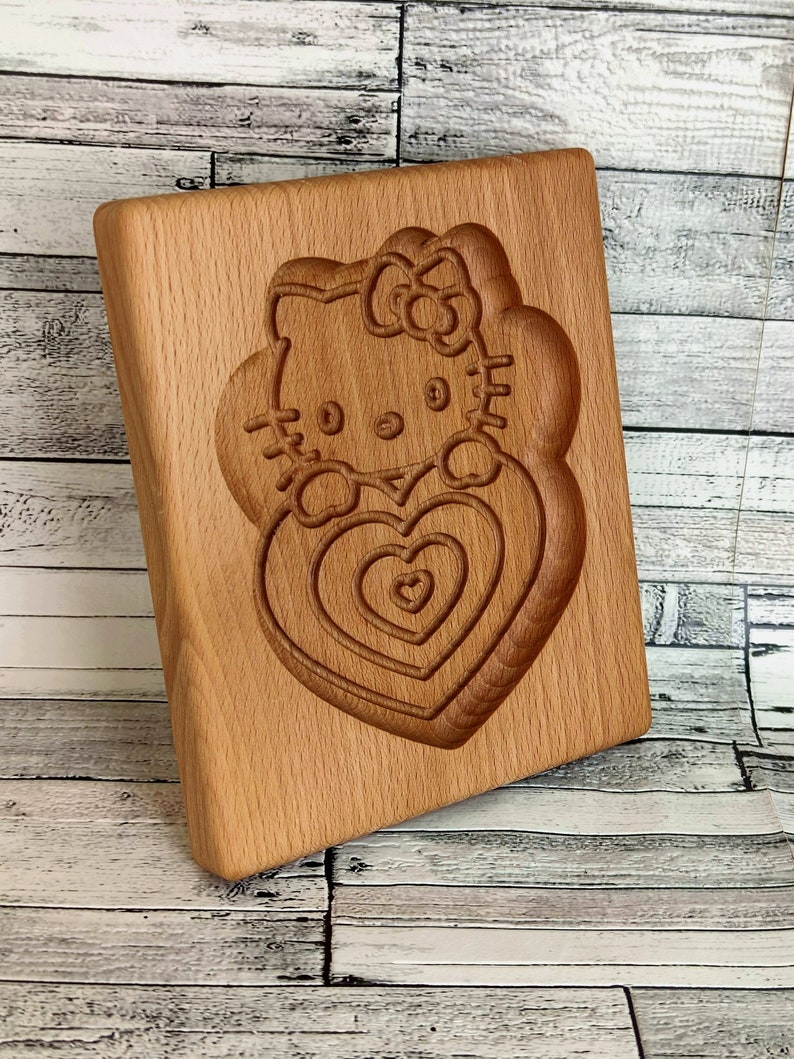 Puede incluir: Cortador de galletas de madera con un dise&ntilde;o tallado de Hello Kitty sosteniendo un coraz&oacute;n. El coraz&oacute;n tiene c&iacute;rculos conc&eacute;ntricos de corazones m&aacute;s peque&ntilde;os.