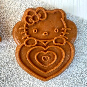 Puede incluir: Una galleta marr&oacute;n con forma de Hello Kitty sosteniendo un coraz&oacute;n. La galleta tiene un contorno detallado de la cara de Hello Kitty y un lazo en la cabeza.