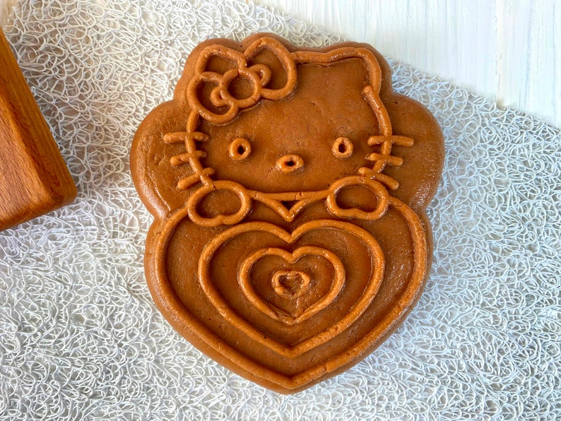 Puede incluir: Una galleta marr&oacute;n con forma de Hello Kitty sosteniendo un coraz&oacute;n. La galleta tiene un contorno detallado de la cara de Hello Kitty y un lazo en la cabeza.