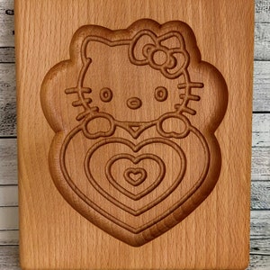 Puede incluir: Una tabla de cortar de madera con un dise&ntilde;o tallado de Hello Kitty sosteniendo un coraz&oacute;n. El coraz&oacute;n tiene c&iacute;rculos conc&eacute;ntricos de corazones m&aacute;s peque&ntilde;os.