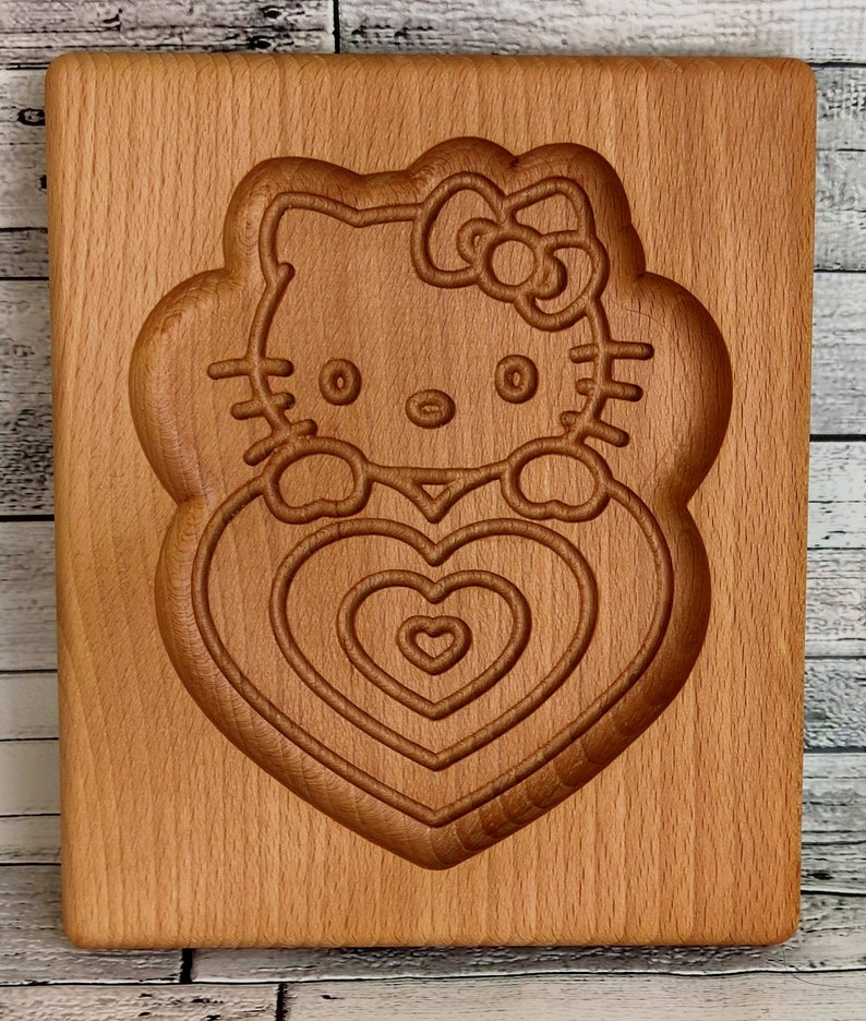 Puede incluir: Una tabla de cortar de madera con un dise&ntilde;o tallado de Hello Kitty sosteniendo un coraz&oacute;n. El coraz&oacute;n tiene c&iacute;rculos conc&eacute;ntricos de corazones m&aacute;s peque&ntilde;os.