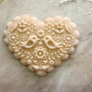 May include: A heart-shaped cookie with a scalloped edge, featuring an embossed design of two birds facing each other, surrounded by floral and botanical patterns. The surface of the light beige dough is lightly dusted with white flour.