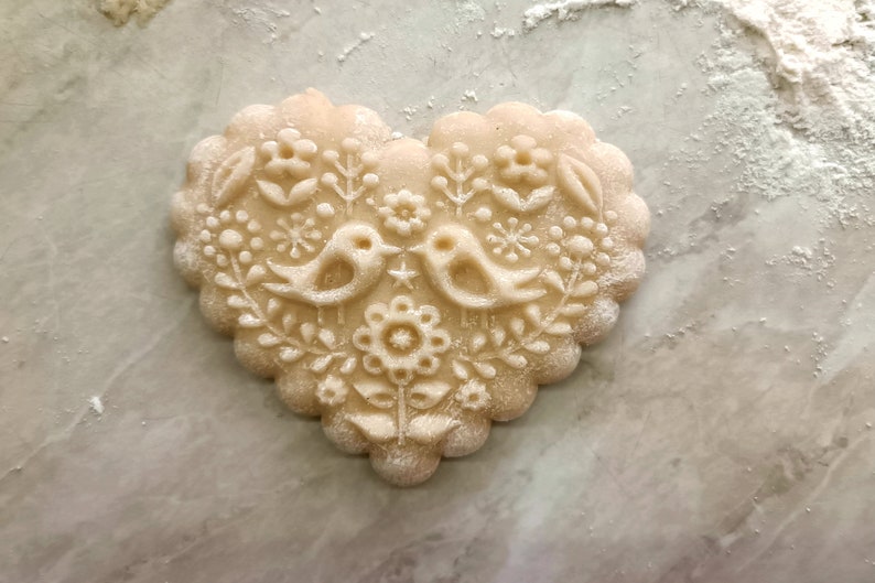 May include: A heart-shaped cookie with a scalloped edge, featuring an embossed design of two birds facing each other, surrounded by floral and botanical patterns. The surface of the light beige dough is lightly dusted with white flour.