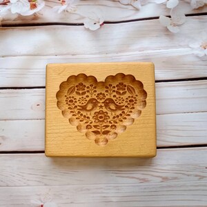 May include: A rectangular light-colored wooden cookie mold featuring a deeply carved heart-shaped design. The center of the heart displays two birds facing each other, surrounded by intricate floral patterns and small star details within a scalloped border.