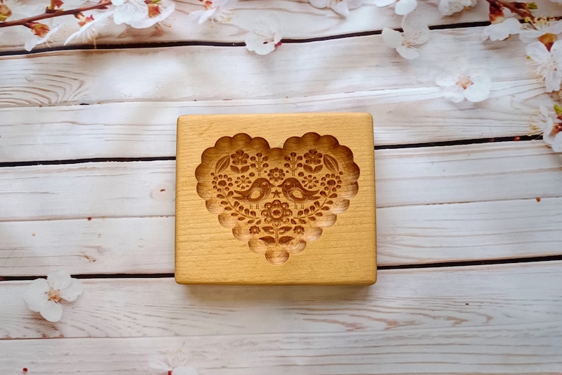 May include: A rectangular light-colored wooden cookie mold featuring a deeply carved heart-shaped design. The center of the heart displays two birds facing each other, surrounded by intricate floral patterns and small star details within a scalloped border.