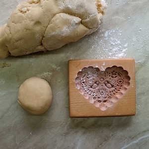 May include: A rectangular wooden cookie mold featuring a carved heart shape with intricate floral patterns and two birds in the center, positioned next to a ball of beige dough and a larger piece of raw dough dusted with white flour.