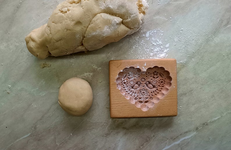 May include: A rectangular wooden cookie mold featuring a carved heart shape with intricate floral patterns and two birds in the center, positioned next to a ball of beige dough and a larger piece of raw dough dusted with white flour.