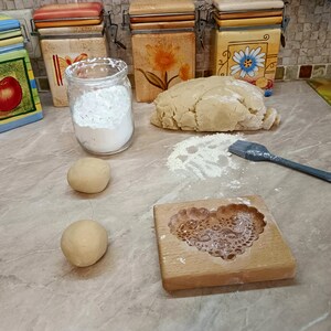 May include: A wooden cookie mold with a heart-shaped floral pattern, two small dough balls, a large mound of dough, a glass jar filled with white flour, and a grey silicone pastry brush. The scene is set on a light brown marble-patterned surface.