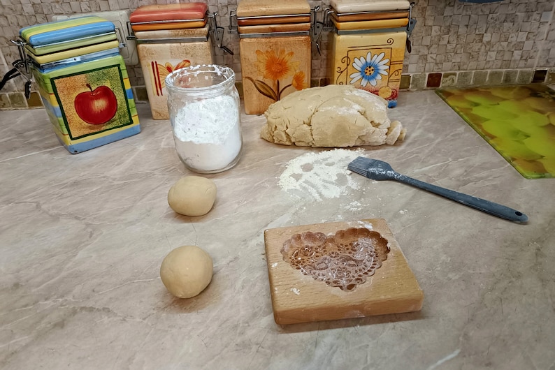 May include: A wooden cookie mold with a heart-shaped floral pattern, two small dough balls, a large mound of dough, a glass jar filled with white flour, and a grey silicone pastry brush. The scene is set on a light brown marble-patterned surface.