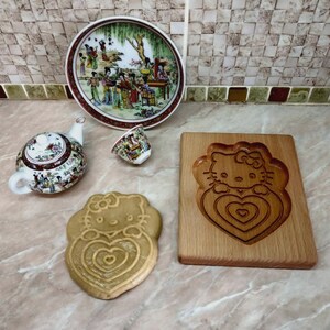 Puede incluir: Una tabla de cortar de madera con un dise&ntilde;o de Hello Kitty tallado. La tabla de cortar es de color marr&oacute;n claro y tiene un acabado de madera natural. Hay una peque&ntilde;a taza de t&eacute; blanca y una tetera con un dise&ntilde;o floral en la superficie de la tabla de cortar.