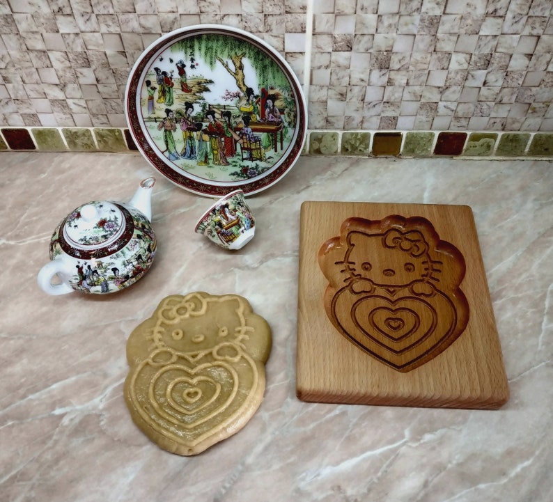 Puede incluir: Una tabla de cortar de madera con un dise&ntilde;o de Hello Kitty tallado. La tabla de cortar es de color marr&oacute;n claro y tiene un acabado de madera natural. Hay una peque&ntilde;a taza de t&eacute; blanca y una tetera con un dise&ntilde;o floral en la superficie de la tabla de cortar.