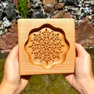May include: A wooden mold with a carved mandala design. The mold is made of light brown wood and has a square shape. The mandala design is intricate and detailed, with a variety of shapes and patterns.