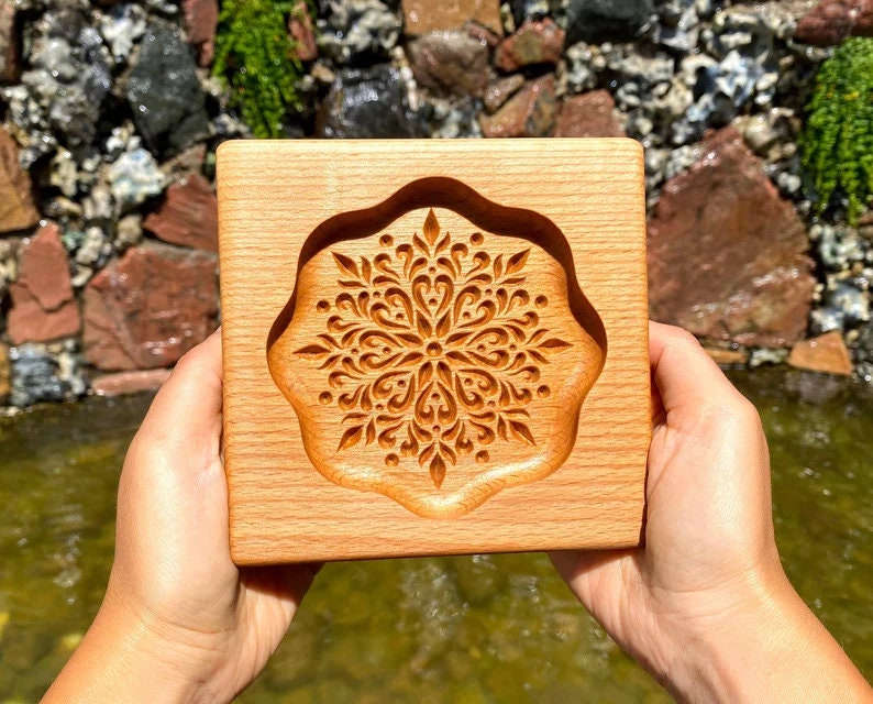 May include: A wooden mold with a carved mandala design. The mold is made of light brown wood and has a square shape. The mandala design is intricate and detailed, with a variety of shapes and patterns.