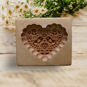 May include: A rectangular light brown wooden cookie mold featuring a deeply carved heart shape. The interior of the heart displays an intricate relief pattern of two facing birds surrounded by various floral and botanical elements.