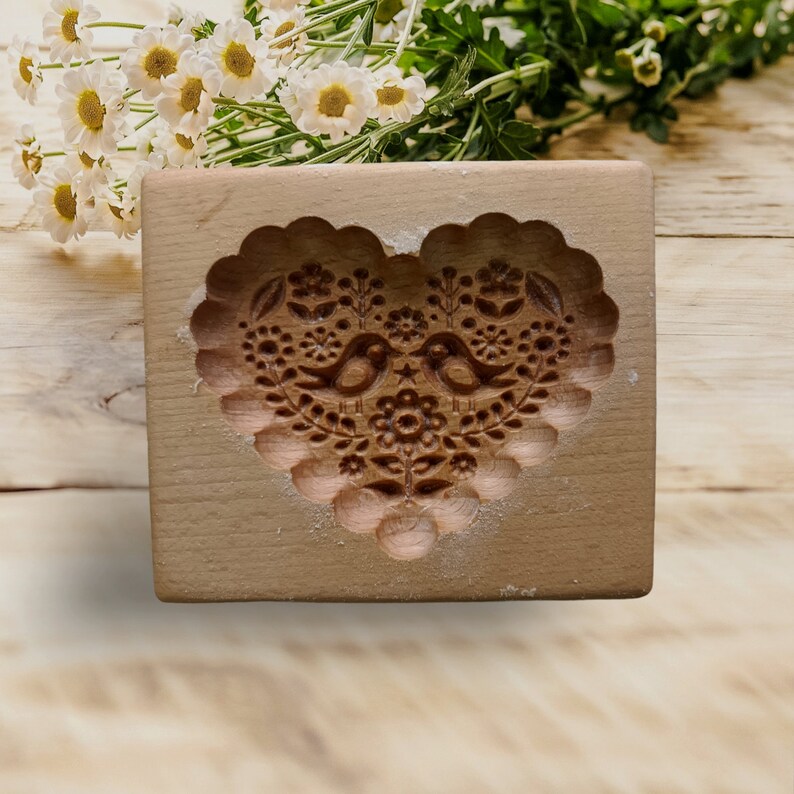 May include: A rectangular light brown wooden cookie mold featuring a deeply carved heart shape. The interior of the heart displays an intricate relief pattern of two facing birds surrounded by various floral and botanical elements.